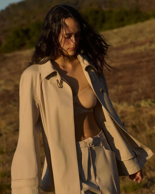 A young adult woman with dark hair wearing a beige jacket and pantsuit stands outdoors in a tan, grassy field with hills in the distance.