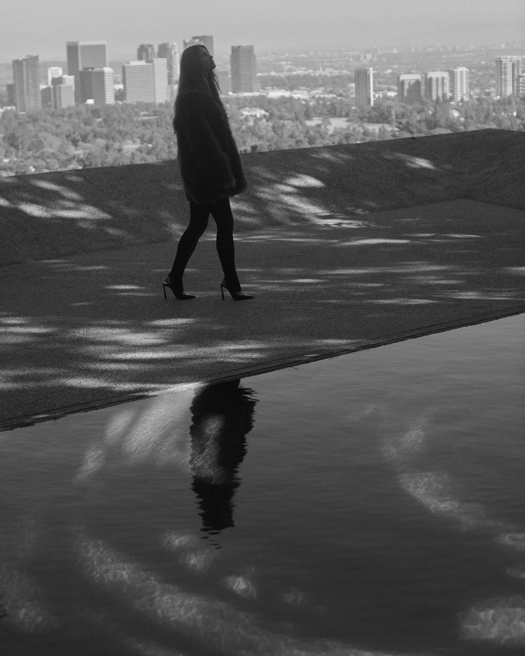 Woman walking on a rooftop terrace overlooking a city skyline with a dark reflection in a pool.