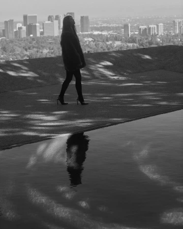 Woman walking on a rooftop terrace overlooking a city skyline with a dark reflection in a pool.