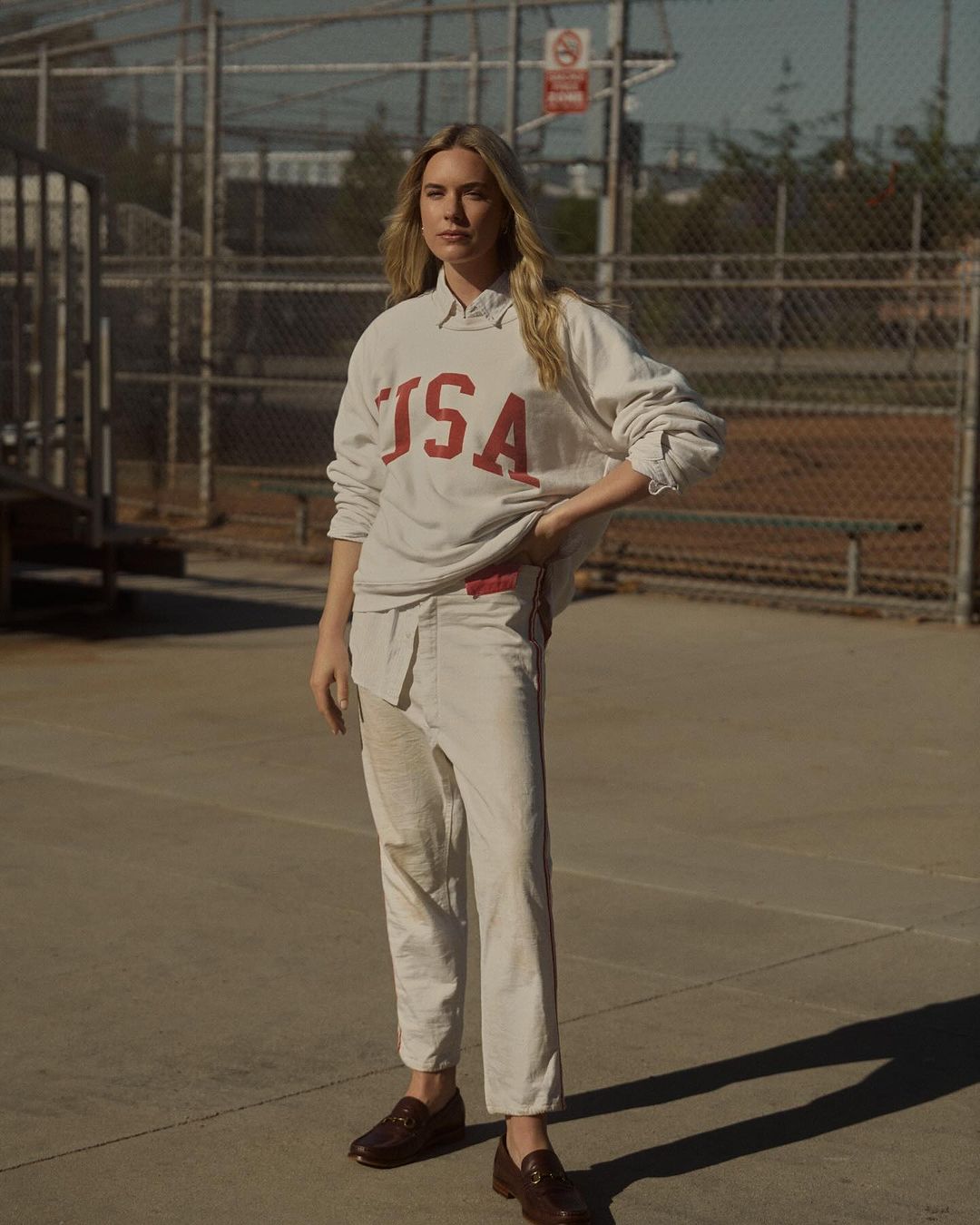 Woman in USA sweatshirt and trousers standing outdoors near a baseball field, posing in full shot, fashion editorial photography.