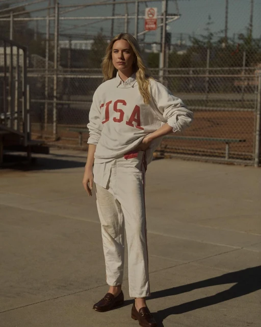 Woman in USA sweatshirt and trousers standing outdoors near a baseball field, posing in full shot, fashion editorial photography.