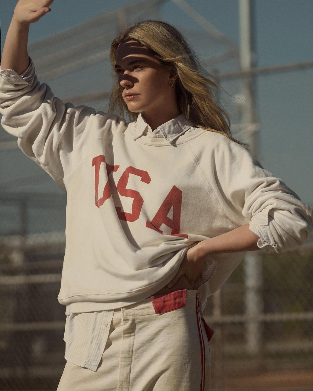A fair-skinned woman in a cream-colored sweatshirt with 'USA' printed on it, stands outside with a relaxed expression and arm raised.
