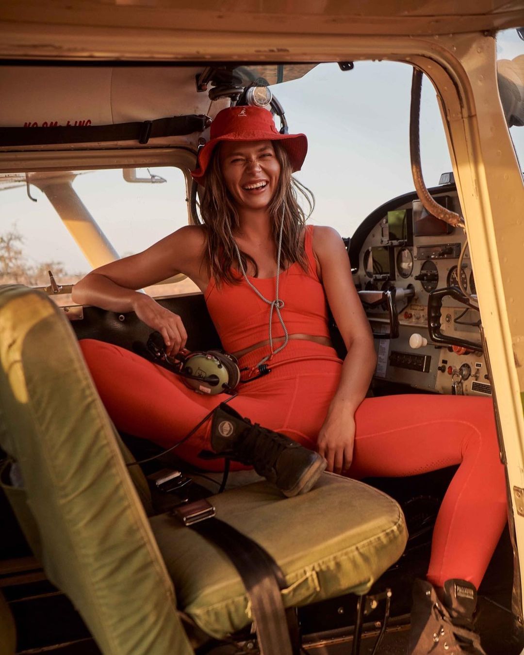 Smiling woman in an airplane cockpit, wearing orange athleisure wear and a hat, accessorized with headset and boots, looking at the camera.