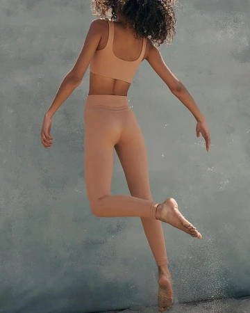 Model with curly hair jumping against a weathered blue wall wearing tan athletic crop top and leggings
