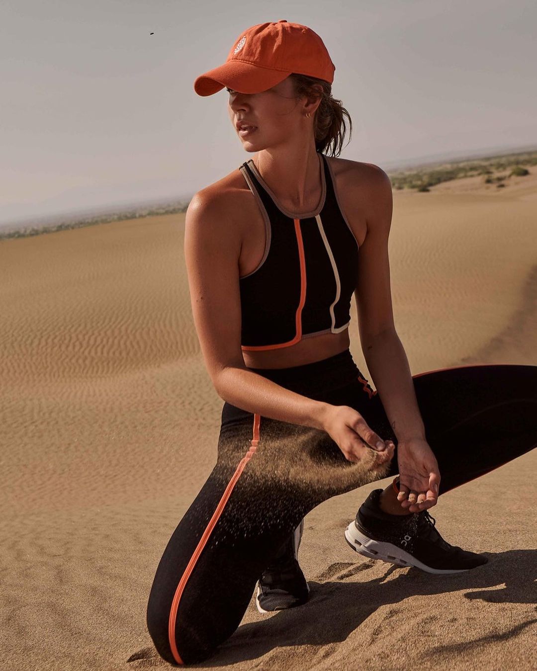 An adult female in athletic wear poses on a sand dune with an orange hat, black top, black leggings, and black athletic shoes.
