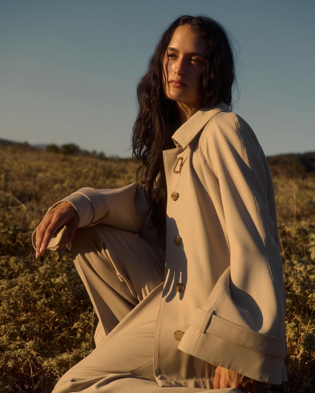 A young adult woman with long dark hair is crouching outdoors, wearing a tan-colored jacket and pants in a lifestyle fashion shot.
