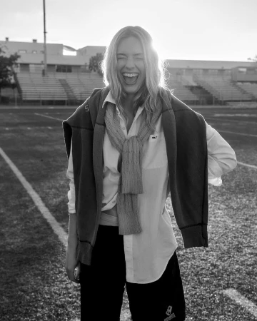 Blonde woman laughing on a sports field wearing a white shirt and sweater tied around her shoulders