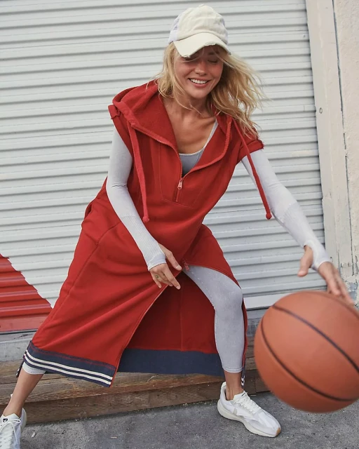 A woman dribbles a basketball on an outdoor court, wearing a rust-colored hoodie dress, white hat, and sneakers, in a sporty lifestyle shot.