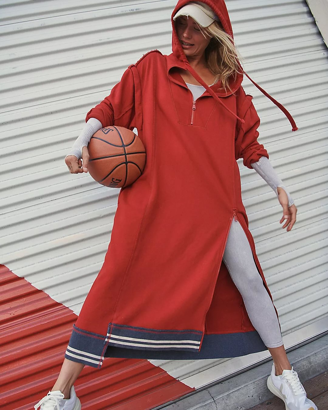 Woman in a red hooded dress holding a basketball in front of a textured wall, wearing white sneakers and a baseball cap.