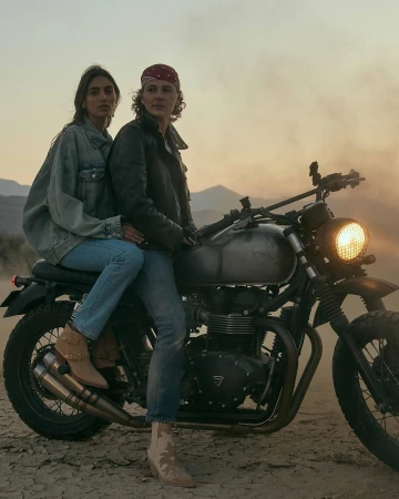 Two people seated on a vintage motorcycle in a dusty landscape at sunset with mountains visible in the hazy background.