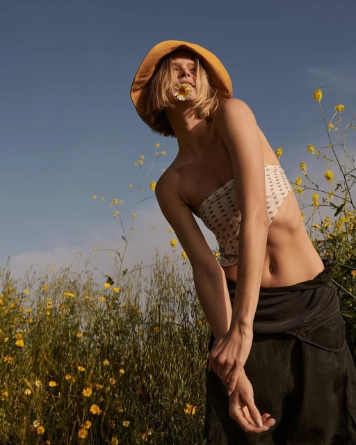 Eye-level portrait of a young woman in a bralette and skirt posing in a field of yellow wildflowers against a sunny blue sky.