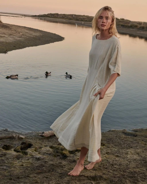 Adult female in neutral dress standing near lake with ducks during golden hour. Fashion and lifestyle photography.