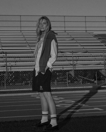 Monochrome portrait of a woman standing on a running track in front of tiered stadium seating, wearing shorts, socks, loafers, and a sweater tied over her shoulders.