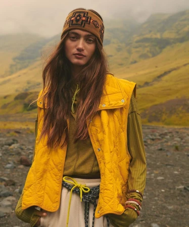 Female fashion model in a quilted vest and patterned headband poses in a rocky, mountainous landscape with misty weather and neutral tones.