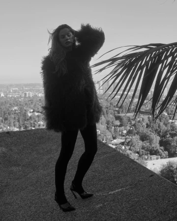 Monochrome portrait of a woman in a textured faux fur coat and high heels posing on a concrete rooftop overlooking a sprawling city