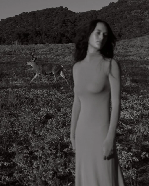 Monochrome image featuring a blurry woman in a dress in the foreground and a deer running across a grassy field, with dark hills in the background.