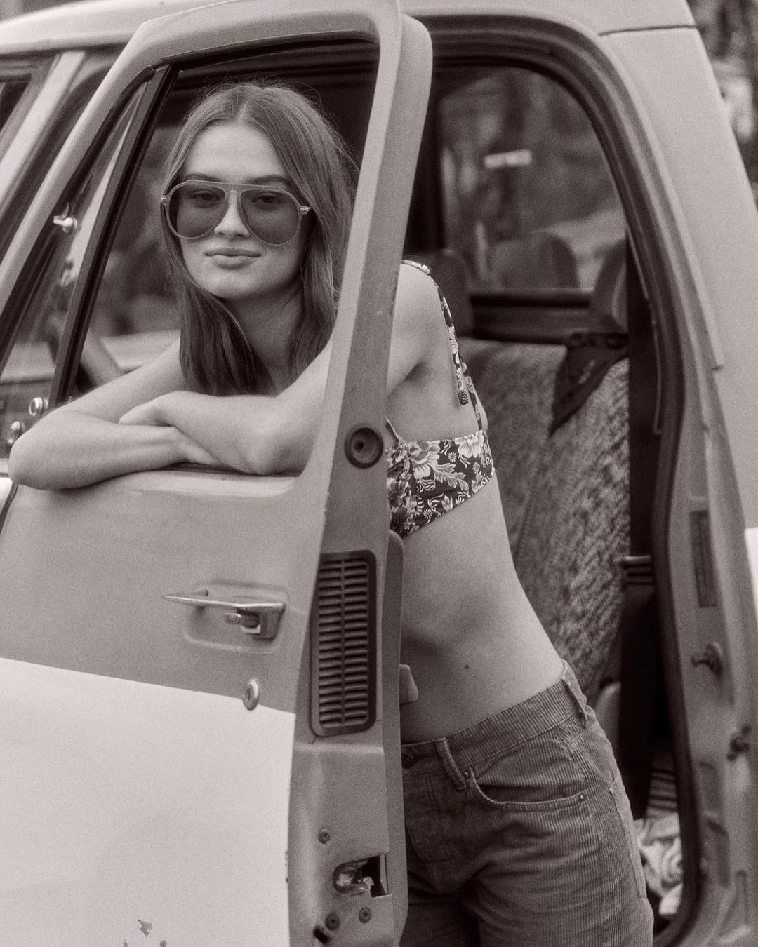 A young woman in sunglasses leans against an open truck door wearing a patterned top and jeans in a high-contrast black and white photograph.
