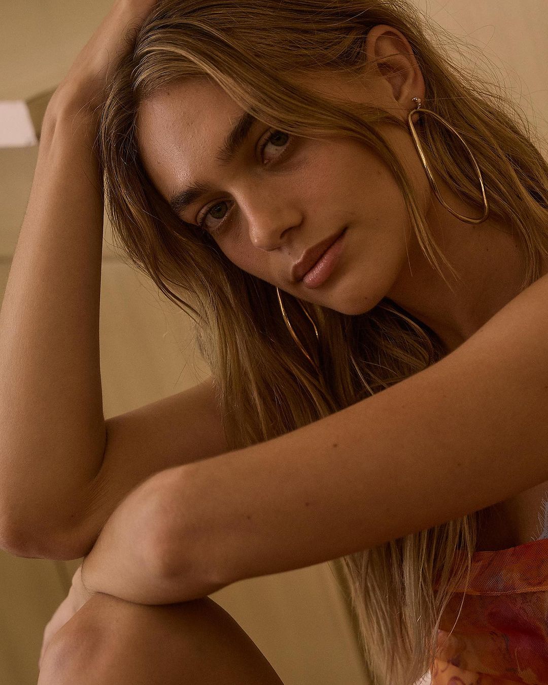 Close-up portrait of a woman with long blonde hair wearing large hoop earrings and a patterned top against a neutral background.