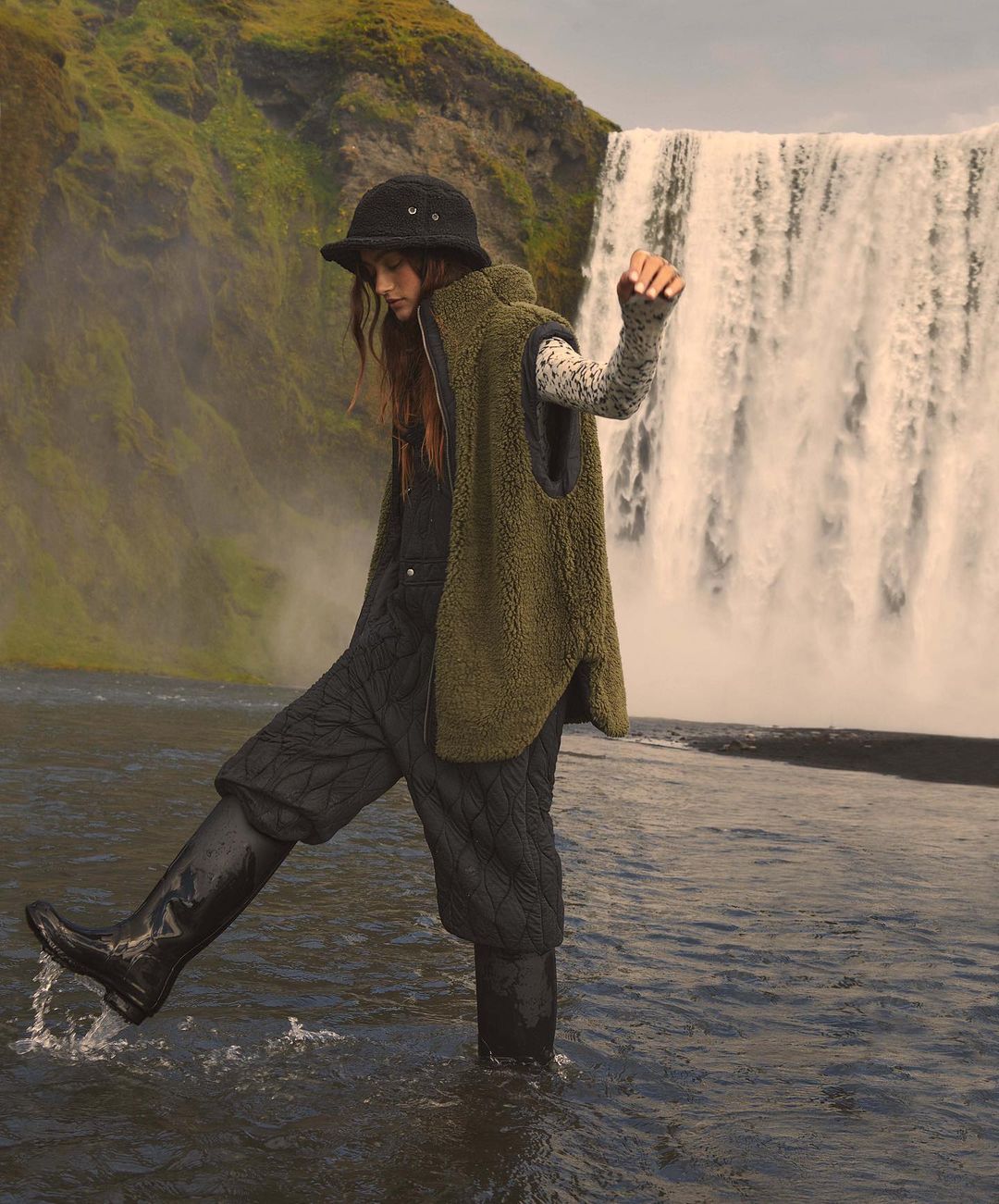 Adult female walks through shallow river near large waterfall wearing a hat, vest, long sleeve shirt, and high rubber boots.