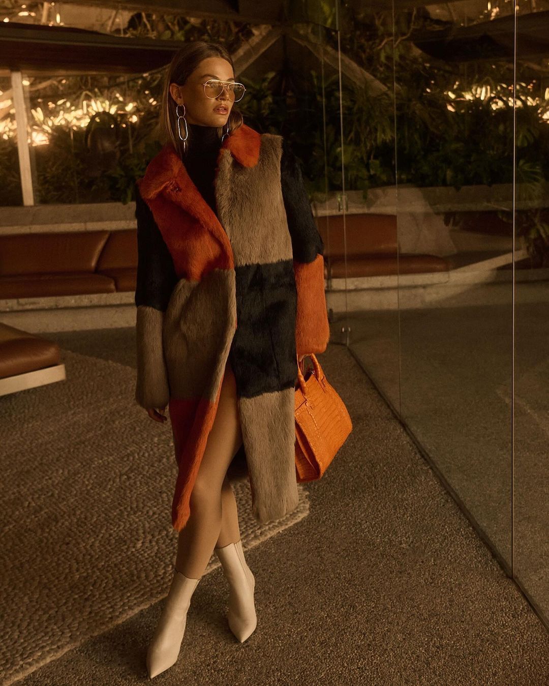 Stylish woman in a color-blocked fur coat, black turtleneck, white boots, and an orange purse poses in an indoor setting with dark lighting.