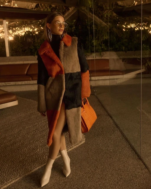 Stylish woman in a color-blocked fur coat, black turtleneck, white boots, and an orange purse poses in an indoor setting with dark lighting.