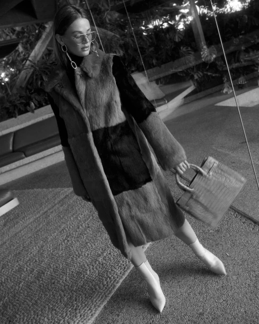 Black and white shot of woman in patchwork coat, turtleneck, boots, and carrying a handbag, posing indoors with foliage.