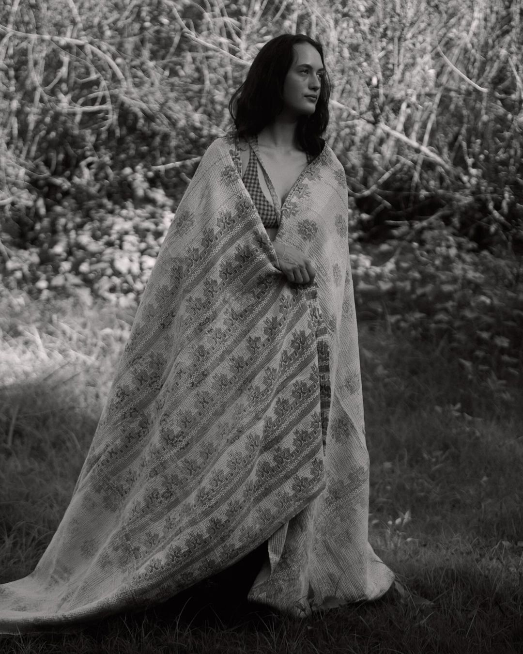 Monochrome portrait of a young adult woman standing in a grassy field covered with a patterned textile, surrounded by foliage and branches.