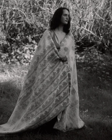 Monochrome portrait of a young adult woman standing in a grassy field covered with a patterned textile, surrounded by foliage and branches.