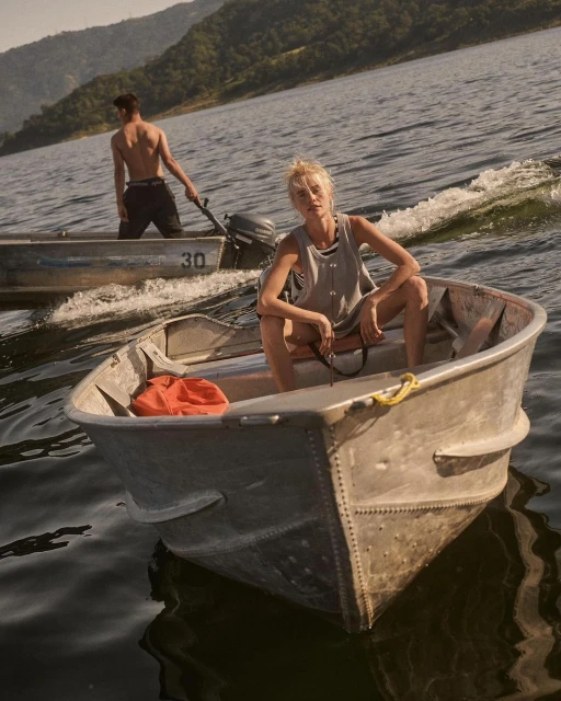 Woman sits in a small boat on a lake with a man in another boat nearby, mountains and trees in the background, creating a serene outdoor scene.