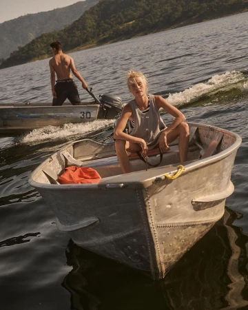 Woman sits in a small boat on a lake with a man in another boat nearby, mountains and trees in the background, creating a serene outdoor scene.