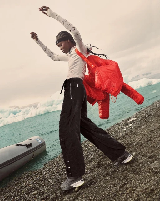Woman in sportswear on black pebble beach by aqua glacial lake with iceberg fragments, wearing red puffer coat and dark sneakers.