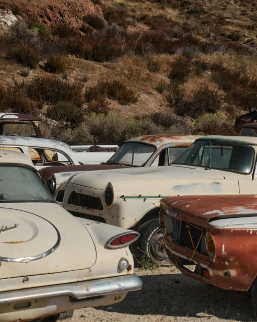 Vintage cars in a junkyard, featuring weathered vehicles and a sun-baked, mountainous backdrop, evoking nostalgia and decay.