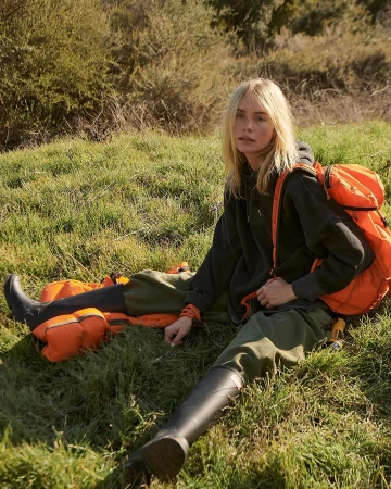 Blonde woman wearing a dark grey hoodie and tall black boots sitting outdoors in a grassy meadow with an orange backpack