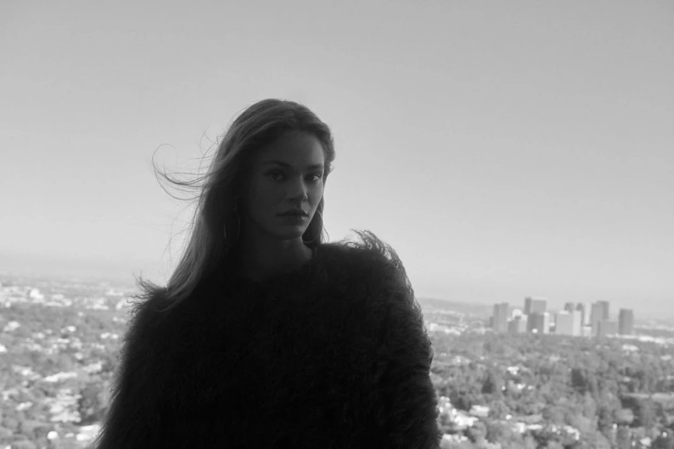 Black-and-white portrait of a woman with long hair wearing a furry jacket, set against a city skyline and a bright sky backdrop.