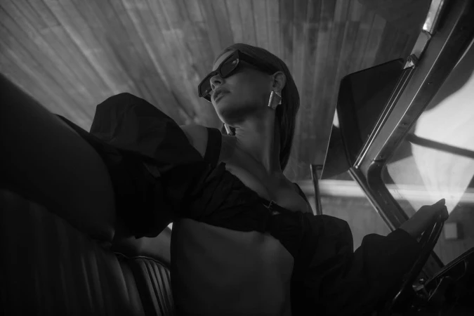 A woman with sunglasses drives a vintage car. She wears a stylish top and geometric earrings in a black and white fashion shot.