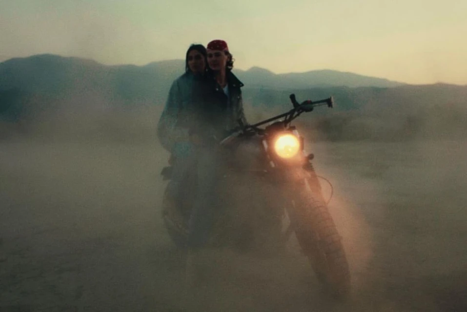 Two people riding a motorcycle through a dusty, desert landscape, silhouetted against a hazy sky with distant mountains.