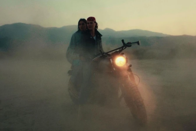 Two people riding a motorcycle through a dusty, desert landscape, silhouetted against a hazy sky with distant mountains.