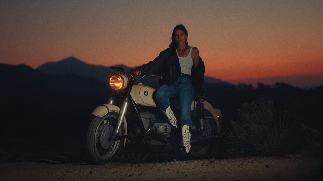Woman in jeans, white tank top, and black jacket sitting on a motorcycle with desert mountains at dusk, wearing white cowboy boots.
