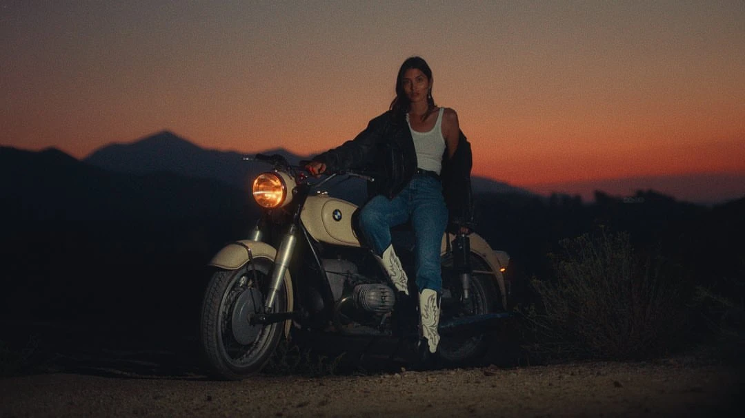 Woman in jeans, white tank top, and black jacket sitting on a motorcycle with desert mountains at dusk, wearing white cowboy boots.
