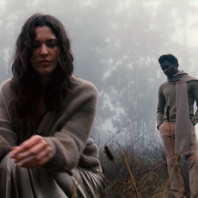 Outdoor fashion shot with a woman crouching in the foreground and a man standing in the misty background, both wearing neutral colors.