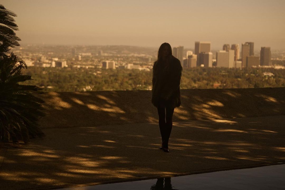 Woman stands silhouetted against an expansive city skyline, bathed in golden light, with a reflecting pool in the foreground.