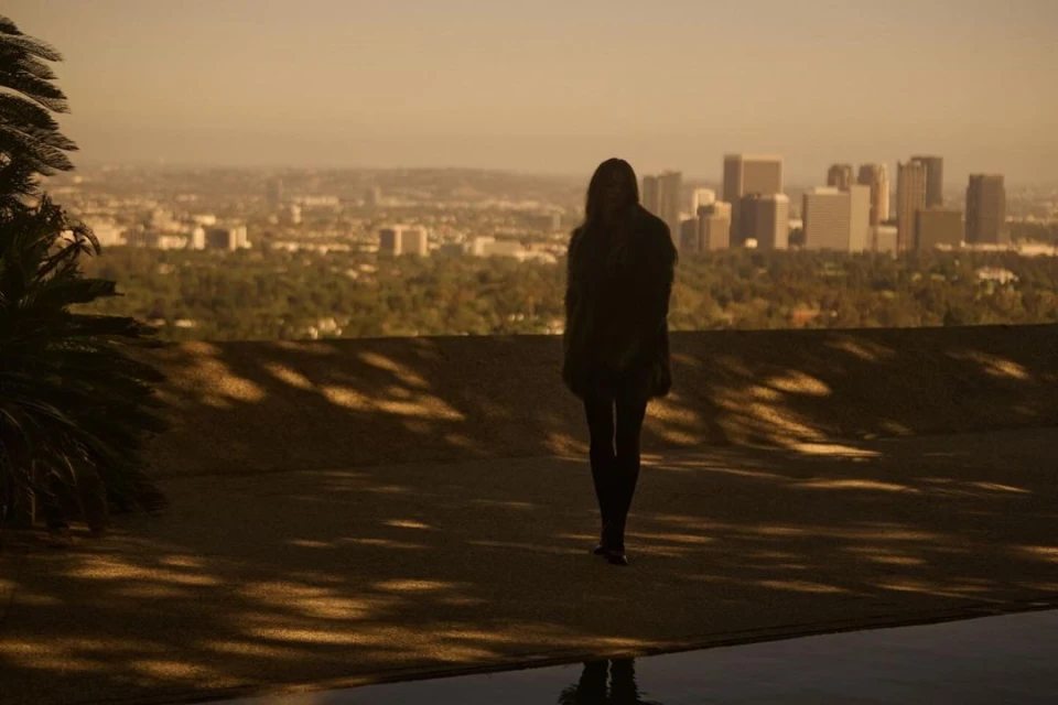 Woman stands silhouetted against an expansive city skyline, bathed in golden light, with a reflecting pool in the foreground.