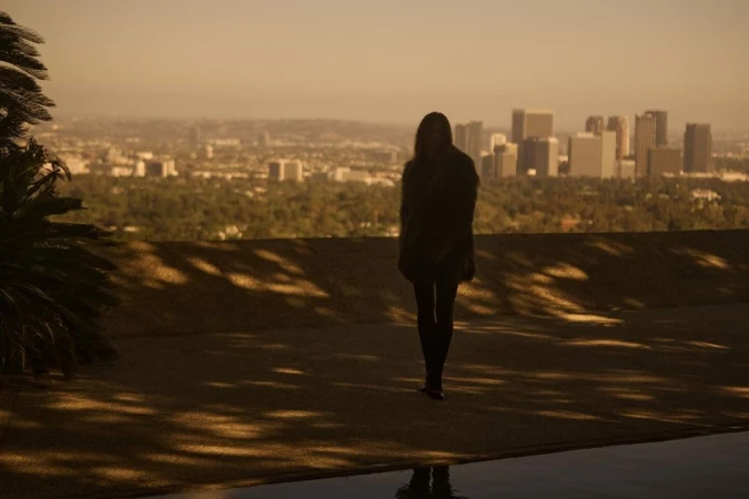 Woman stands silhouetted against an expansive city skyline, bathed in golden light, with a reflecting pool in the foreground.
