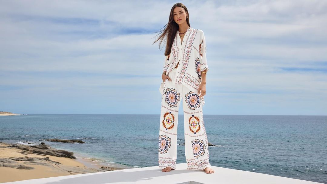 Model wearing patterned white blouse and matching trousers standing outdoors against an ocean and sky background