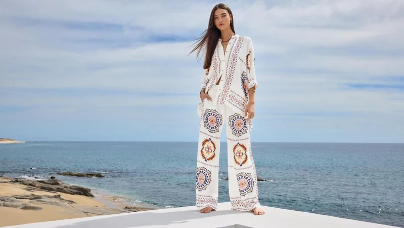 Model wearing patterned white blouse and matching trousers standing outdoors against an ocean and sky background