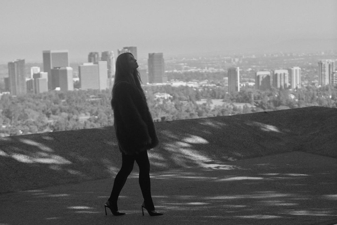 Monochrome full-length shot of a woman in a fur coat walking on a rooftop overlooking a city skyline.