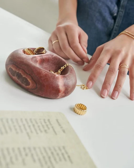 Close-up of hands with rings on, arranging a gold chain necklace near a brown jewelry holder and a gold ring on a white surface.