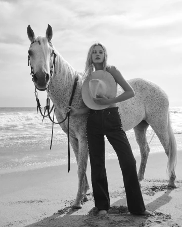 Blonde woman in dark jeans standing on a sandy beach holding a wide-brimmed hat next to a white horse