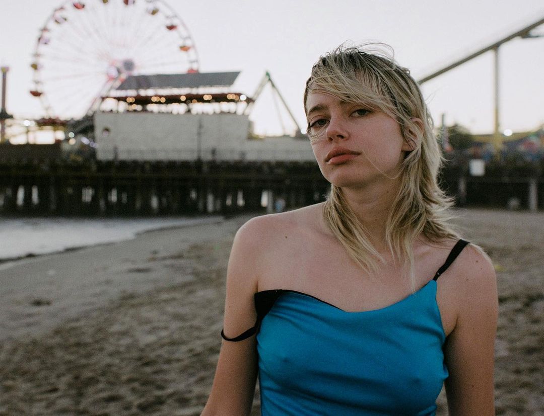 Portrait of a young woman in a blue dress on a sandy beach with a pier and Ferris wheel in the background during daytime.