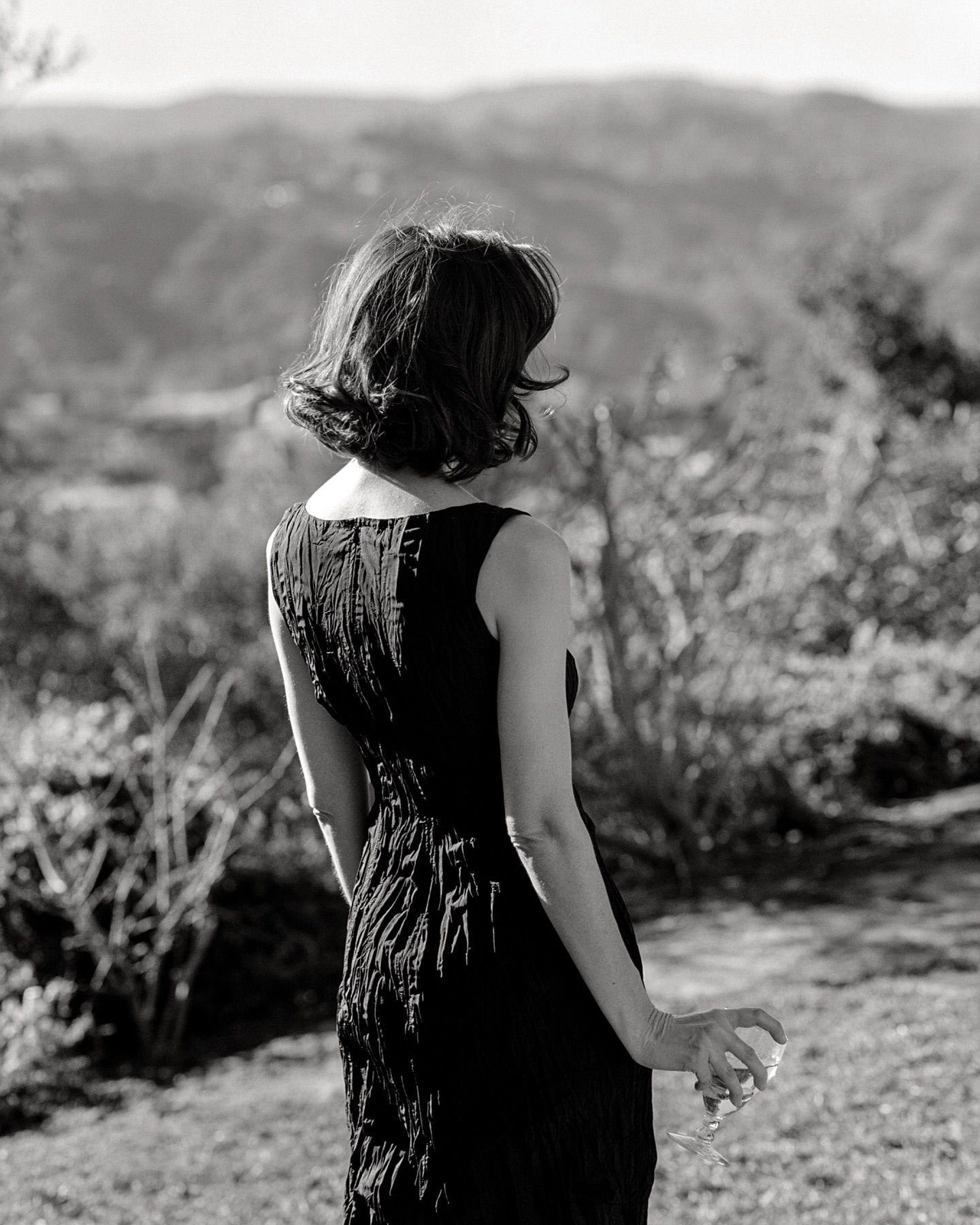 Woman in a black dress holding a wine glass outdoors with hills in the background, captured in monochrome by Chloe Chippendale.